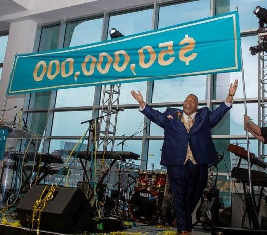 Dr. 迈克尔·巴斯顿，高举双臂，站在舞台上，横幅上宣布5000万美元的目标已经实现